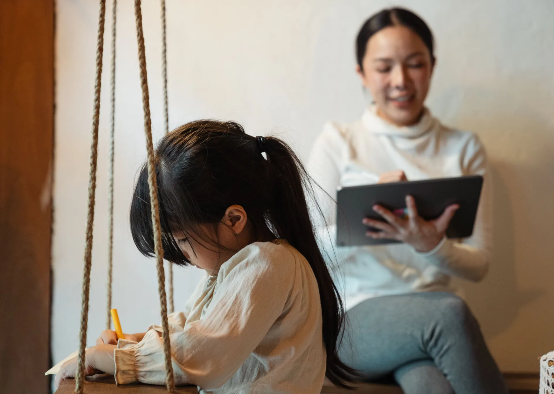 A clinician holding a tablet smiles as she observes a young girl drawing at a table