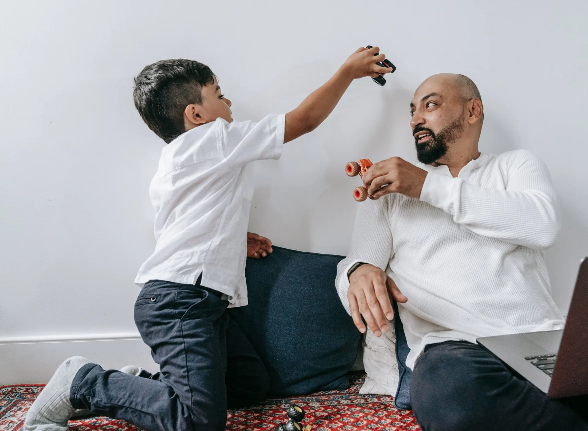 A smiling dad sitting on the living room floor playing with monster trucks alongside his young son